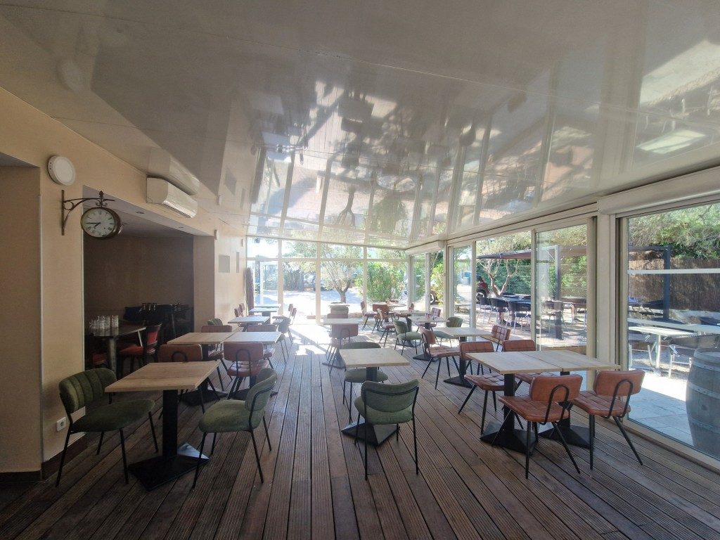 La salle vitrée de la brasserie : tables en bois, chaises vertes et orange, plancher en bois et grandes baies sur la terrasse