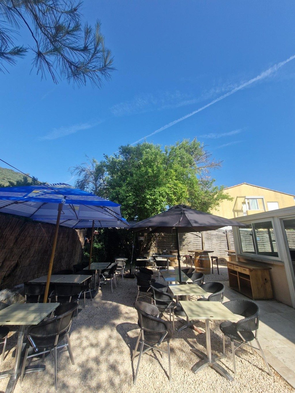 La terrasse du bistrot : tables et chaises sous les arbres et les parasols, sur du gravier